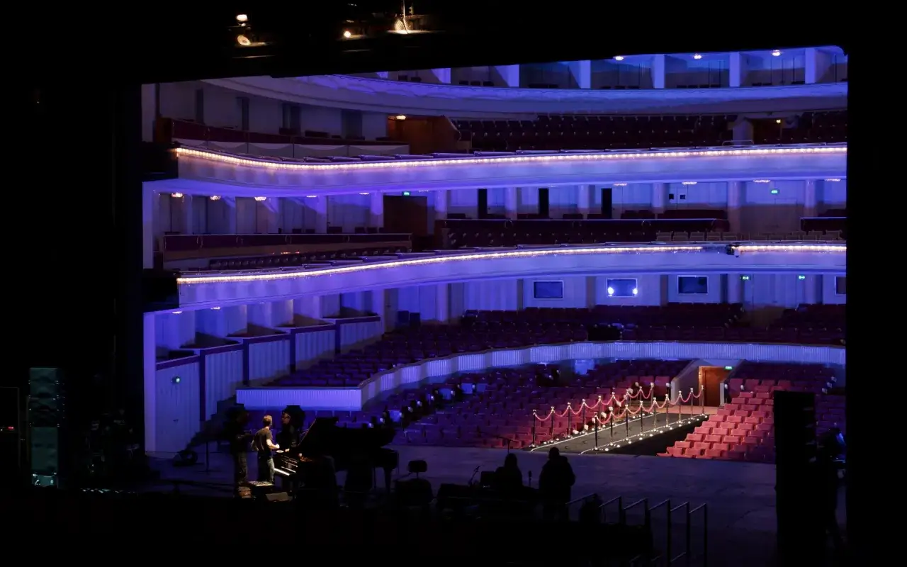Widok na pustą widownię teatru wielkiego, oświetloną niebieskim światłem. Na scenie przygotowania do występu.