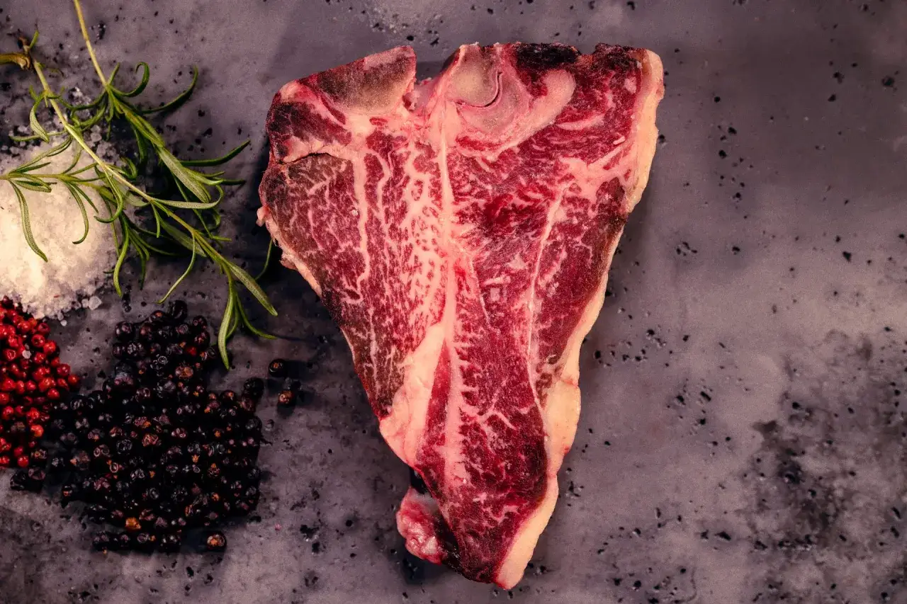 Duży stek T-bone z widocznym marmurkowaniem, gotowy do domowego sezonowania wołowiny na sucho w zwykłej lodówce. Obok sól, pieprz i rozmaryn.