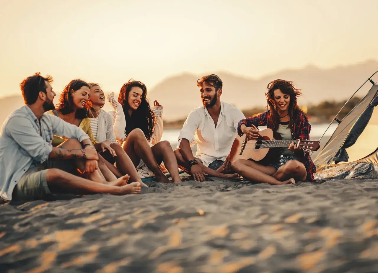 Grupa przyjaci&oacute;ł siedzi na plaży przy namiocie, śmiejąc się i grając na gitarze. Idealne miejsce, by spędzić wakacje w lipcu.