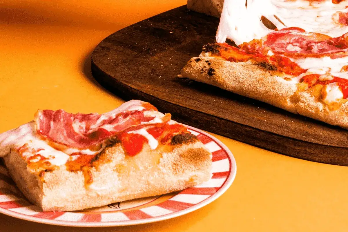 Kawałek pysznej pizzy rzymskiej z szynką i mozzarellą na talerzu. Obok leży reszta pizzy na desce.