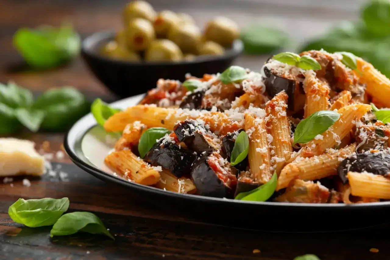 Rigatoni alla norma z bakłażanem, pomidorami i bazylią, posypane parmezanem. Obok miseczka oliwek.