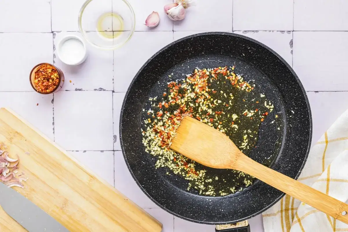 Na patelni smaży się czosnek i chili, przygotowując bazę do spaghetti aglio e olio. Obok leżą składniki: oliwa, sól, posiekana szalotka.