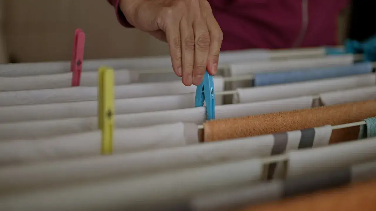 Dłoń z niebieską klamerką wiesza białą koszulę na suszarce. W tle widać inne ubrania i klamerki.