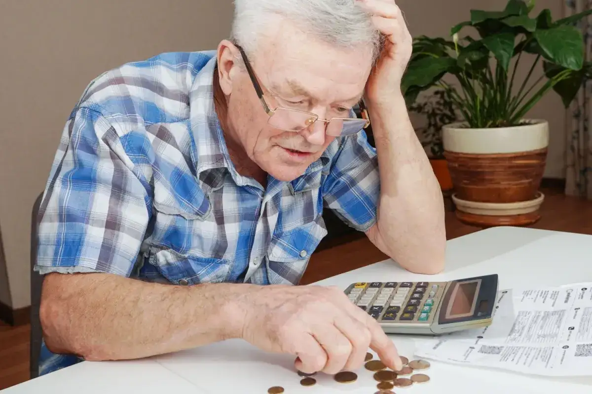 Starszy mężczyzna z kalkulatorem i monetami na stole, analizuje finanse po odprawie emerytalnej w prywatnej firmie.