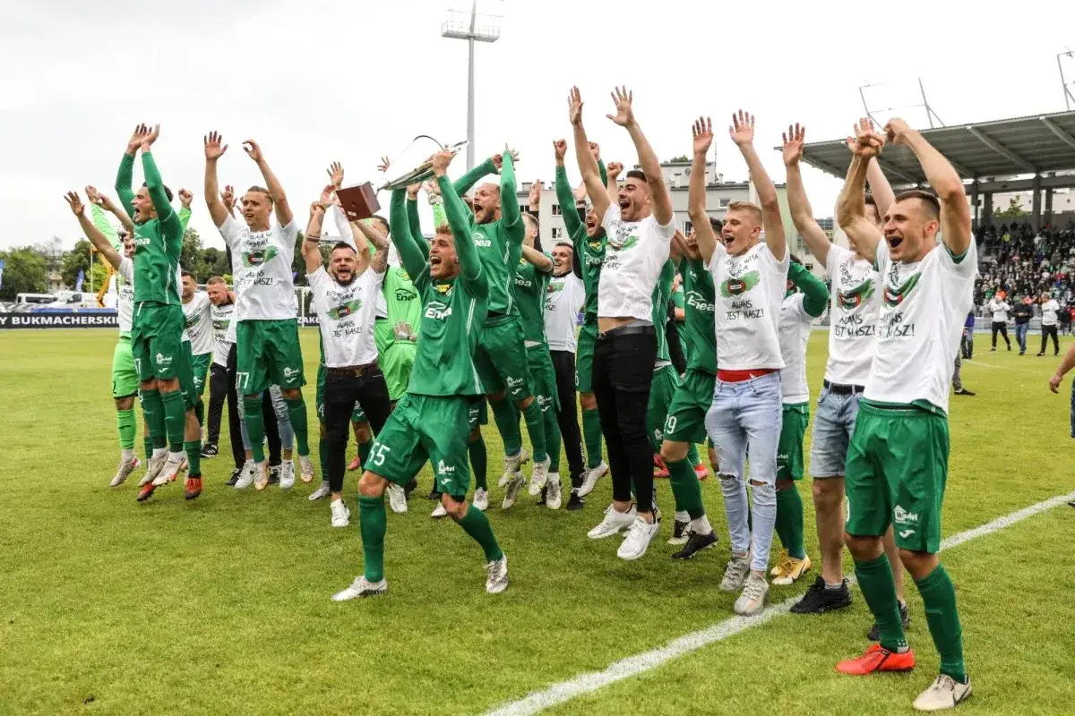 Piłkarze w zielonych strojach świętują, unosząc ręce i puchar. Na koszulkach napis 