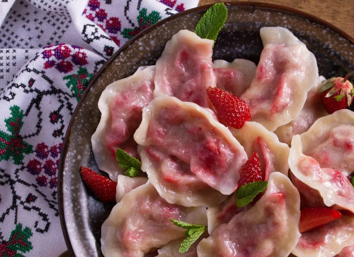 Pyszne pierogi z truskawkami, ozdobione świeżymi owocami i miętą, na tle haftowanej serwetki.