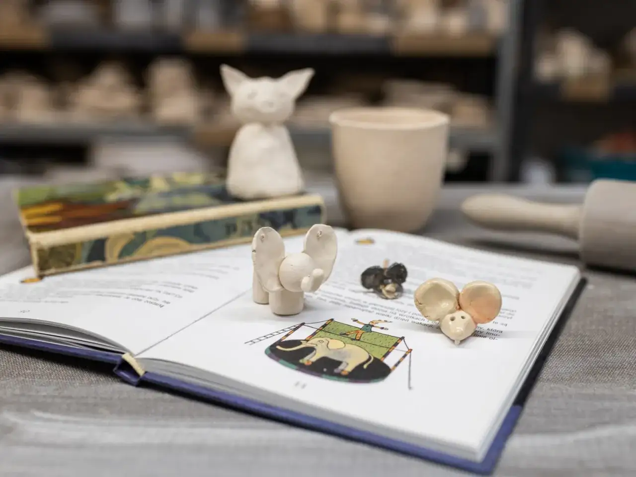 Urocze figurki z gliny, słonik i myszki, ozdabiają otwartą książkę. To efekt kreatywnych warsztat&oacute;w z gliny dla dzieci.