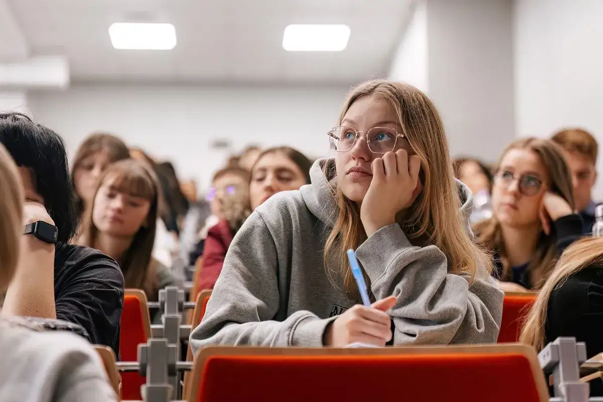 Uczniowie w sali wykładowej, zastanawiający się, czy w technikum jest matura. Dziewczyna w okularach zamyślona, z długopisem w dłoni.