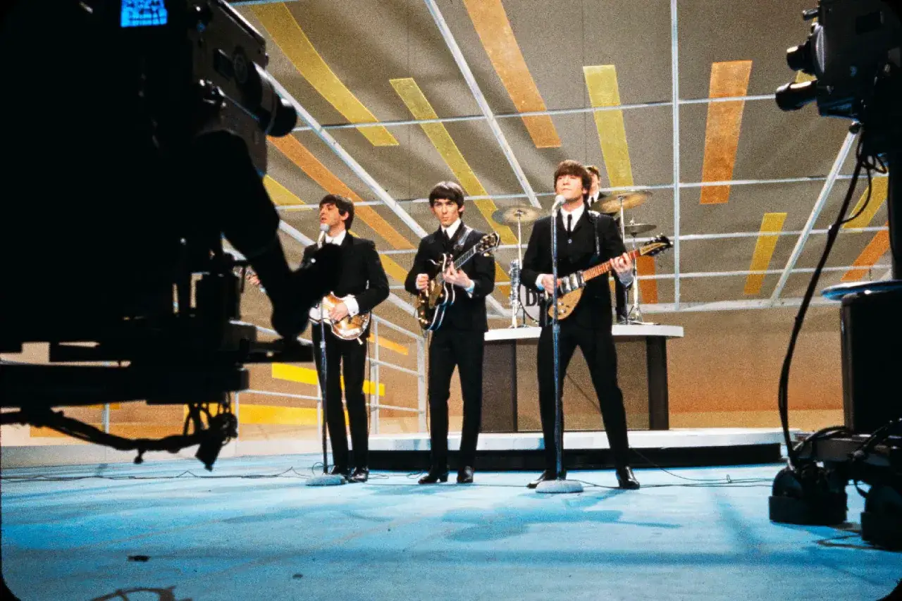 The Beatles w czarnych garniturach grają na gitarach na scenie. Ciekawe ujęcie zza kamery.