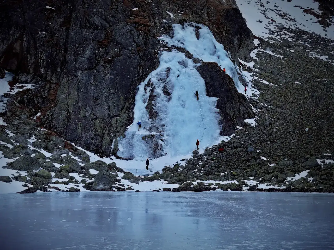 Wspinacze na lodospadzie Kościelca zimą. Lodowy wodospad i skalne zbocza pokryte śniegiem, przed nimi zamarznięte jezioro.