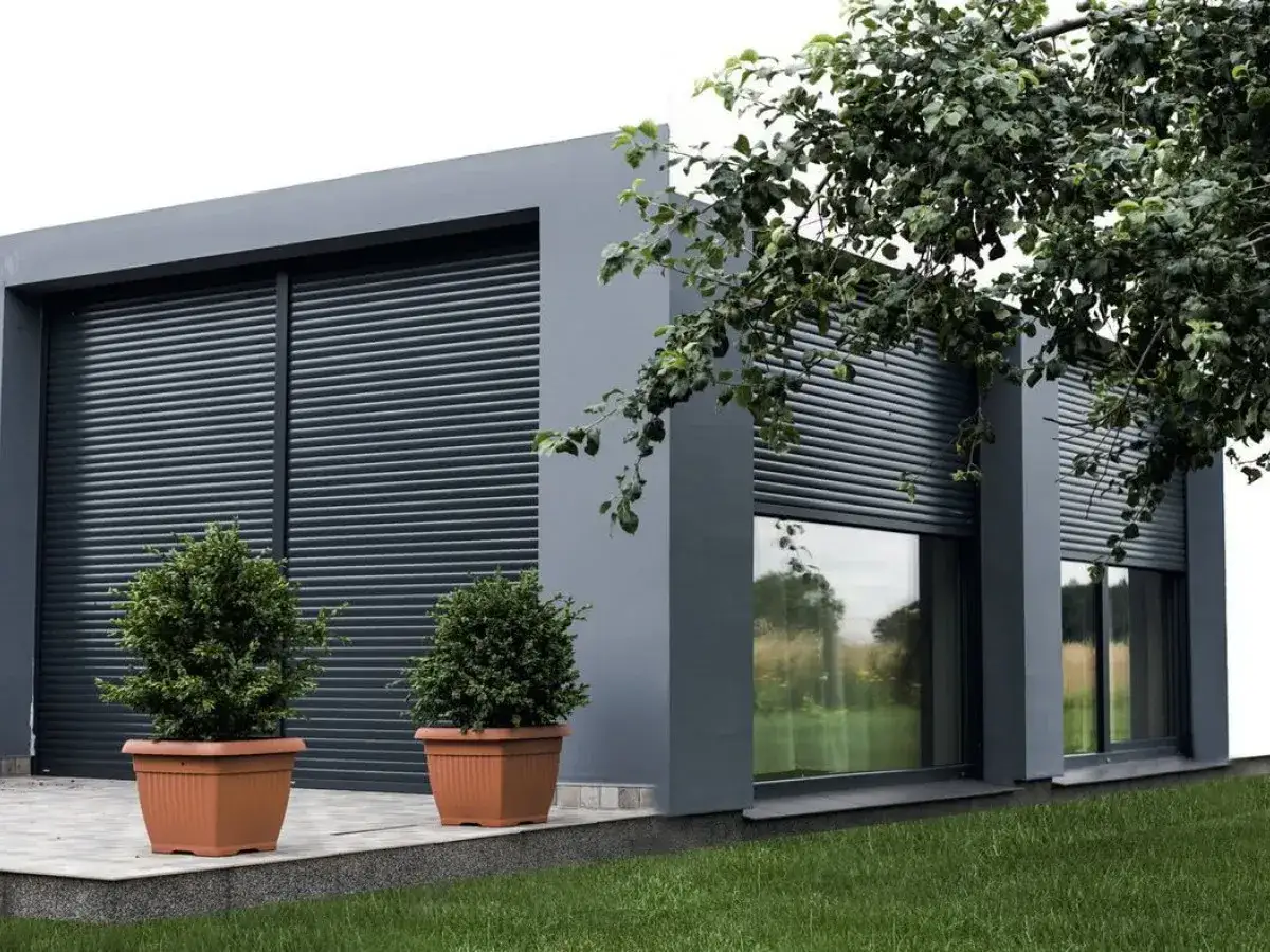 Nowoczesny budynek z szarymi roletami nakładanymi na okna i drzwi. Dwa krzewy w donicach przed wejściem.