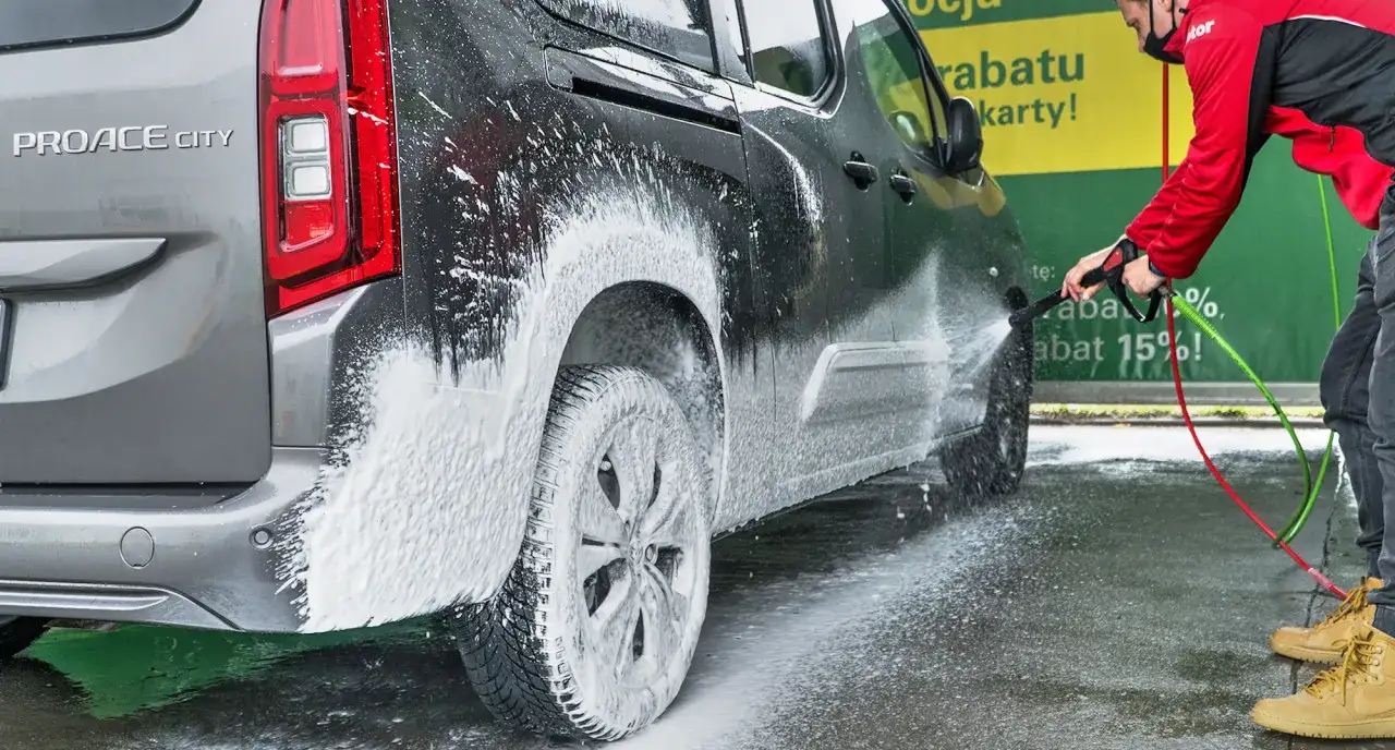 Mężczyzna w czerwonej kurtce pokazuje, jak myć auto na myjni, spryskując je pianą.