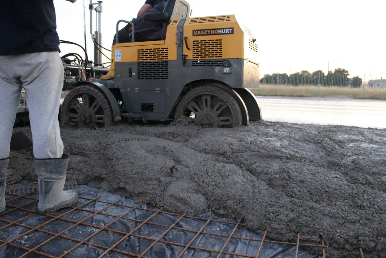 Maszyna budowlana wylewa beton, by czym wyr&oacute;wnać posadzkę. Na pierwszym planie widać zbrojenie i gumowce pracownika.