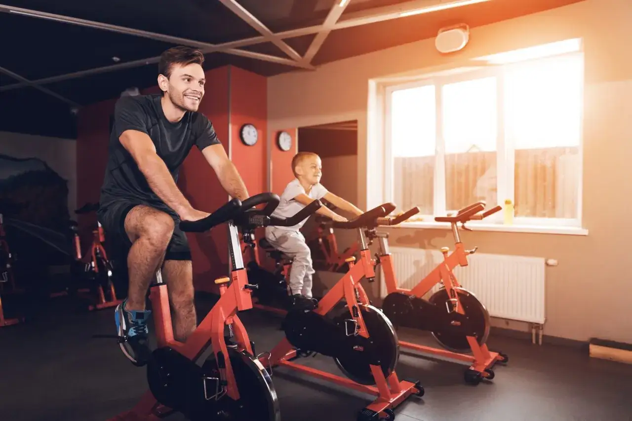 Ojciec i syn cieszą się wspólnym treningiem na rowerze stacjonarnym. Uśmiechnięci, pedałują obok siebie w siłowni.