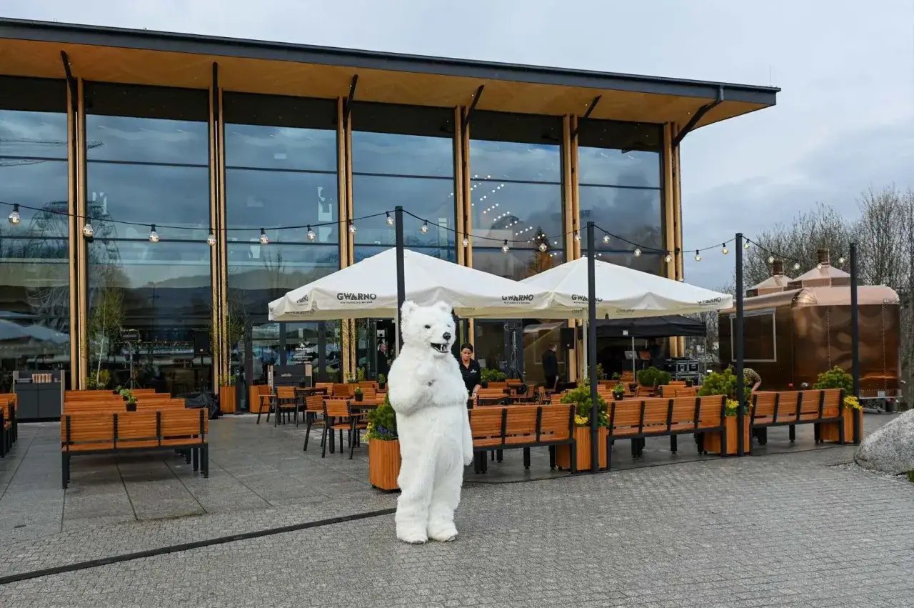 Pluszowy miś wita gości przed nowoczesną karczmą na wierchu. W tle widać ogr&oacute;dek z parasolami i miedziany food truck.