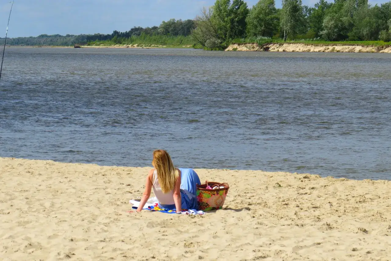 Dziewczyna siedzi na piaszczystej plaży w Ciszycy, patrząc na spokojną rzekę. Obok niej kolorowy koszyk plażowy.