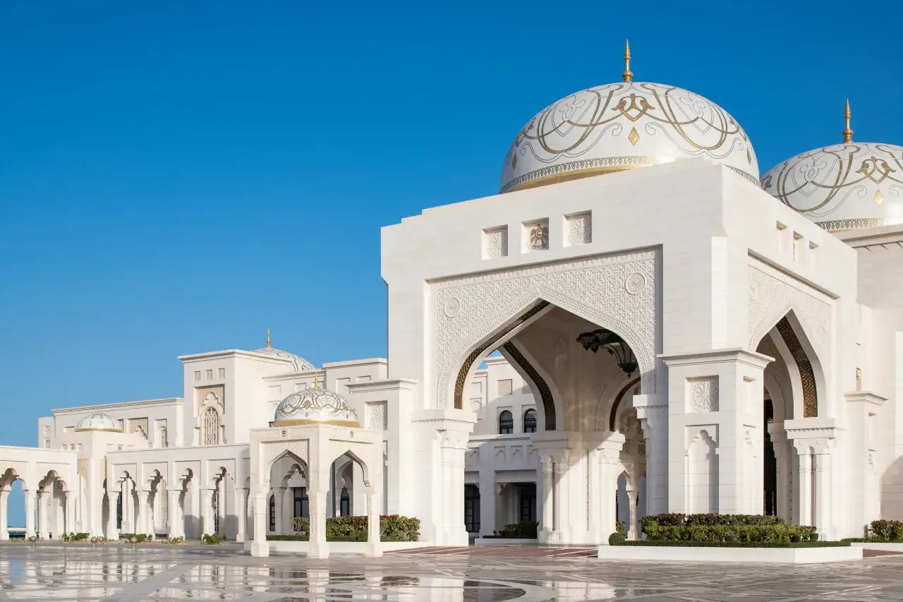 Majestatyczny Qasr Al Watan z białego kamienia, z ozdobnymi kopułami i łukami, odbija się w mokrym placu pod błękitnym niebem.