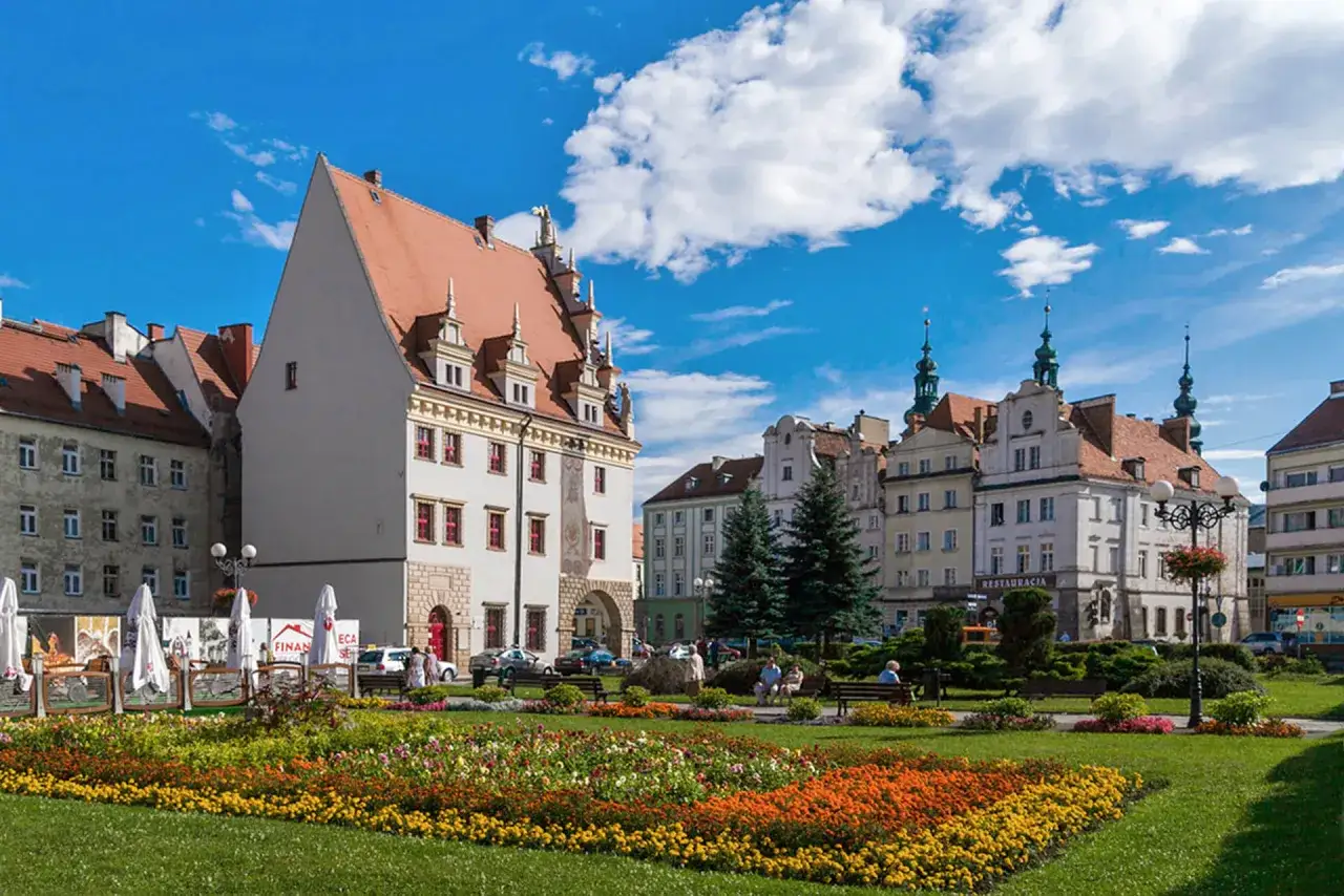 Kolorowy plac w Nysie z kwitnącymi rabatami, zabytkowymi kamienicami i błękitnym niebem. Idealne miejsce na spacer i odkrywanie atrakcji.