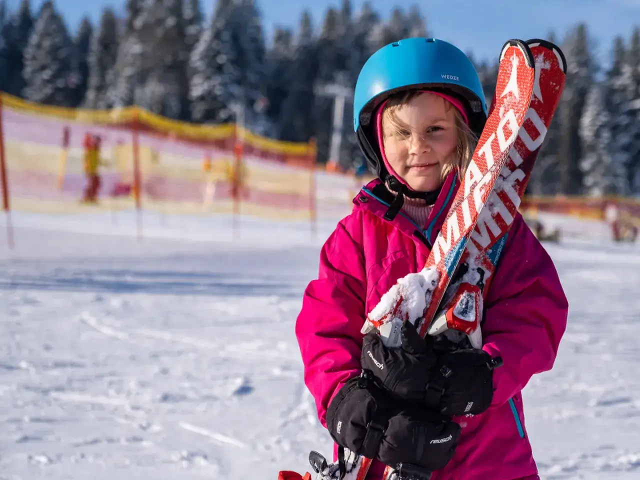 Mała narciarka w różowej kurtce i niebieskim kasku trzyma narty. Idealne miejsce, gdzie na narty do Czech, z widokiem na ośnieżone drzewa.