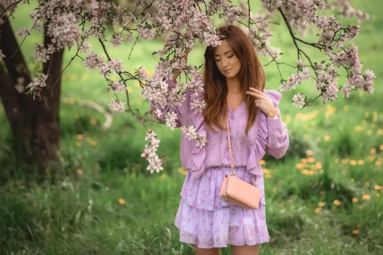 Kobieta w kwiecistej sukience i sweterku w kolorze liliowym, pod kwitnącym drzewem.