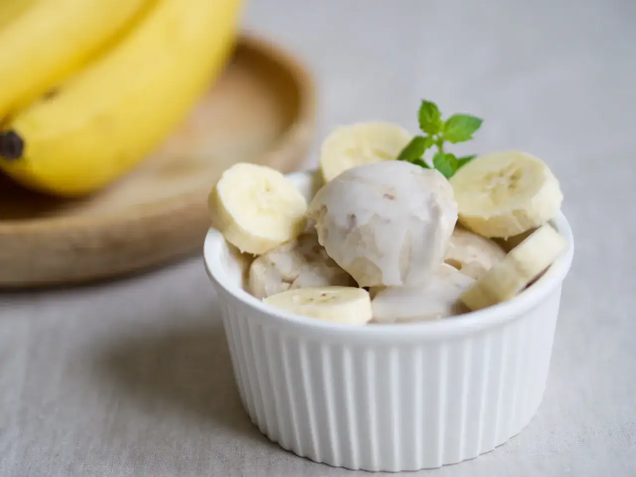 Pyszne lody bananowe w białej miseczce, ozdobione listkiem mięty. Obok leżą świeże banany.