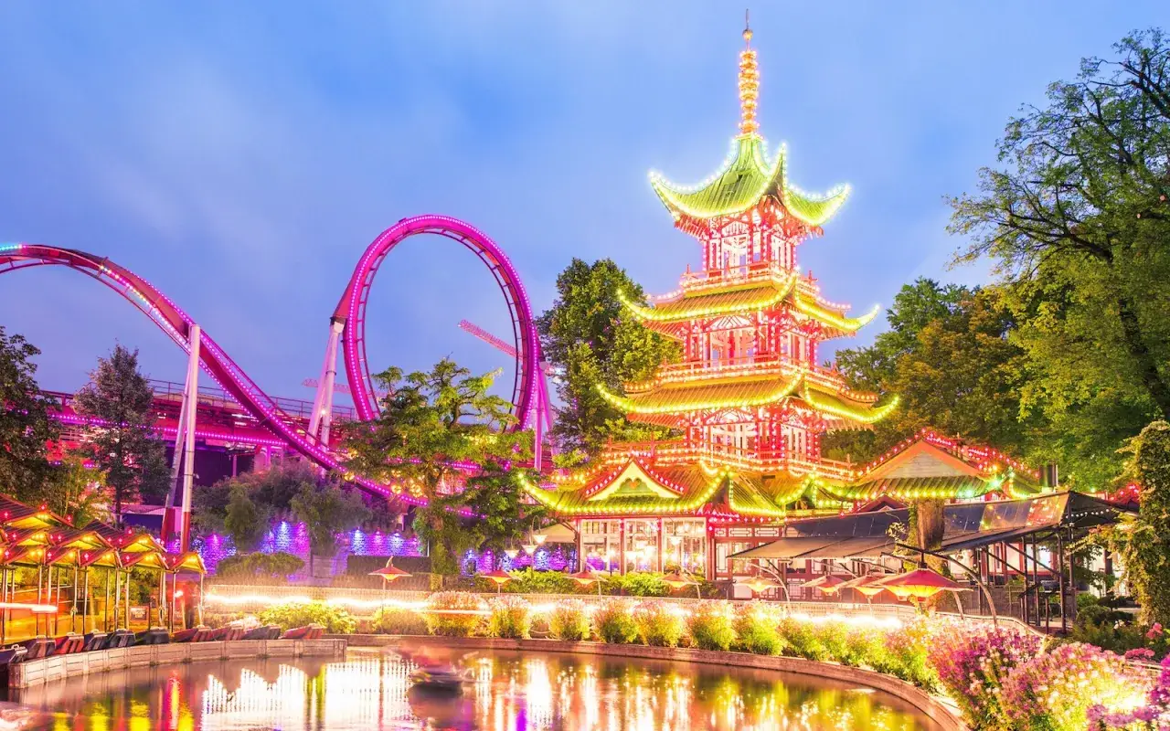 Magiczny wieczór w parku Tivoli Kopenhaga. Pagoda i różowa kolejka górska lśnią w świetle tysięcy lampek.