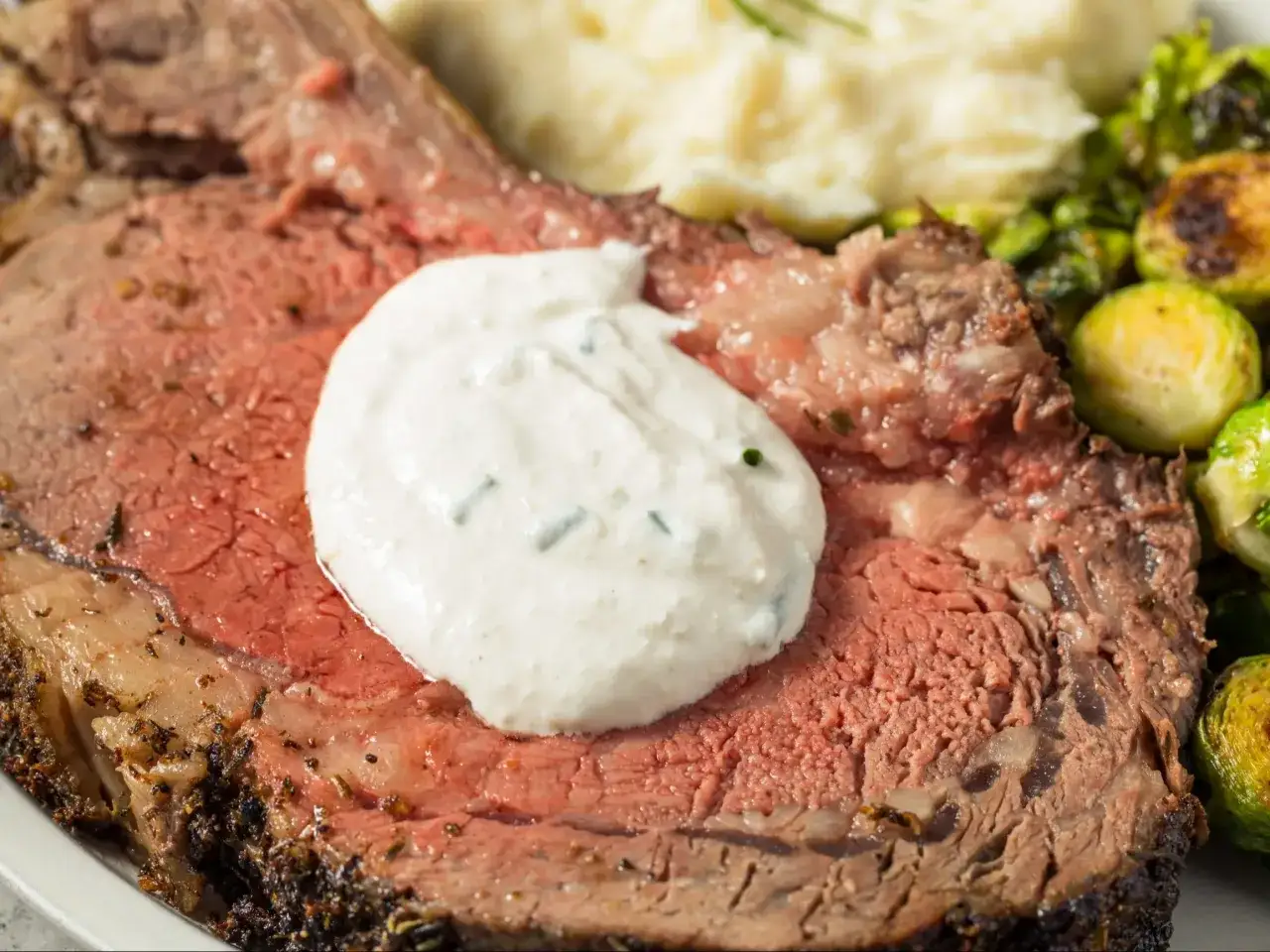 Pyszny pieczony rostbef z kremowym, ciepłym sosem chrzanowym na wierzchu, podany z ziemniakami i brukselką.