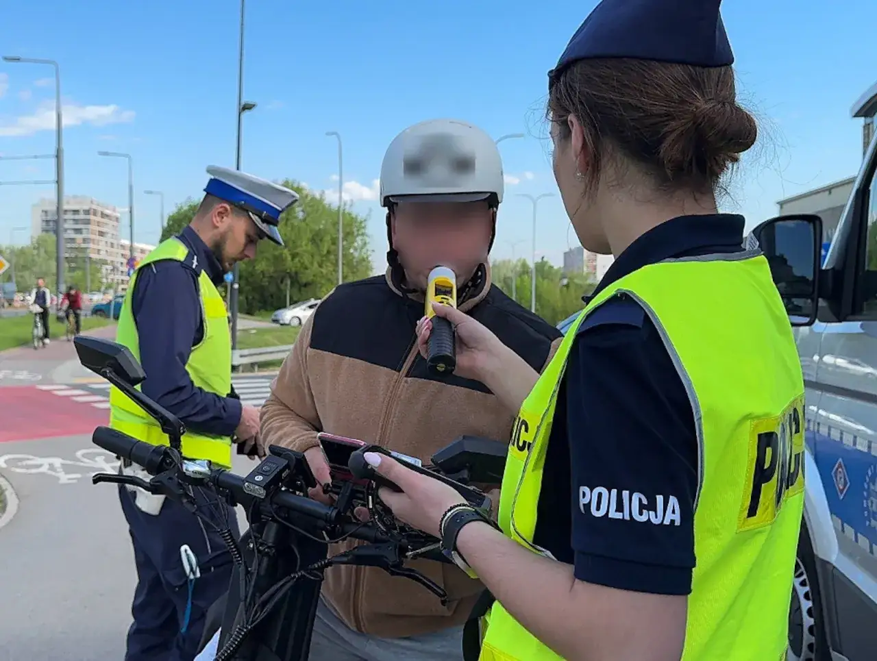 Policjanci sprawdzają, czy można jechać rowerem po alkoholu. Mężczyzna dmucha w alkomat, a funkcjonariuszka z radiowozem obserwuje.