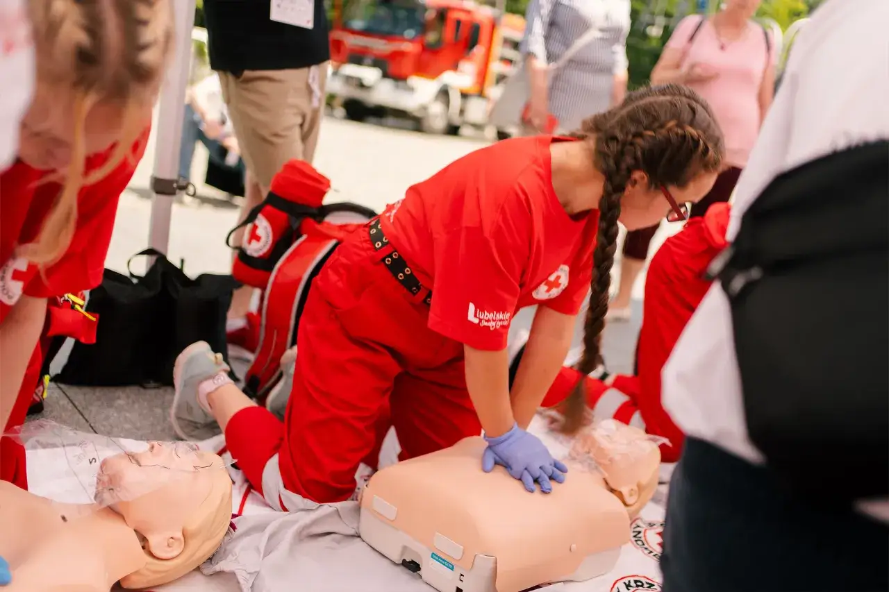 Osoby w czerwonych strojach ćwiczą resuscytację na fantomie. Pokazują, kto może prowadzić kurs pierwszej pomocy.