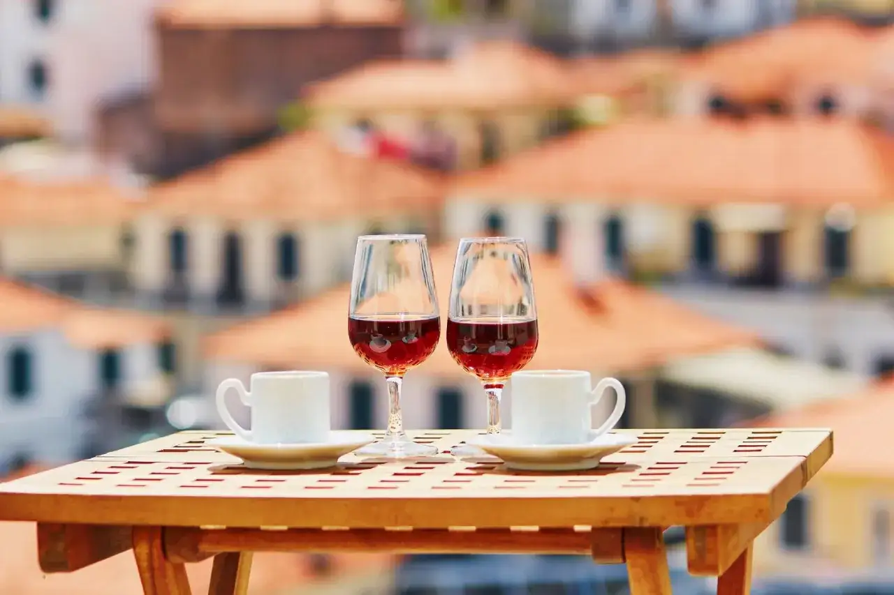 Dwa kieliszki z bursztynowym winem z Madery i dwie filiżanki kawy na drewnianym stoliku z widokiem na dachy miasta.