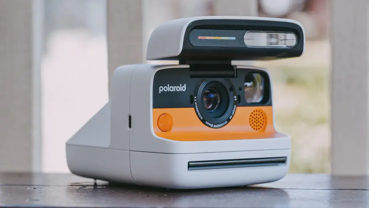 Pomarańczowo-biały aparat fotograficzny który drukuje zdjęcia, z charakterystycznym logo Polaroid i lampą błyskową.