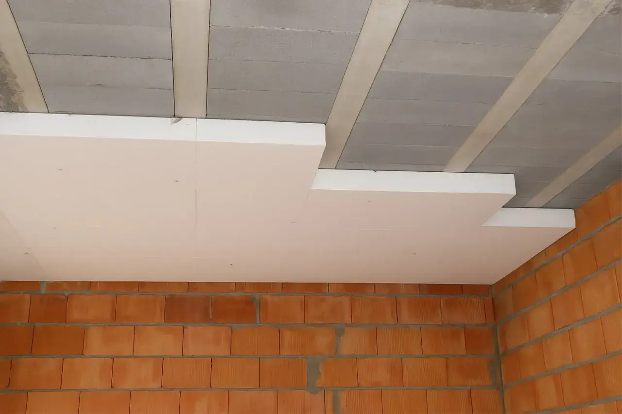 Białe płyty styropianowe przyklejane do betonowego stropu. Ceglane ściany poniżej. Dowiedz się, czym kleić styropian do sufitu.