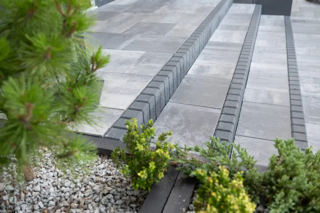 Nowoczesne schody z szarej kostki brukowej, z ozdobnym obrzeżem. W tle zieleń iglaków i kamyczki. Idealny przykład, jaki beton pod kostkę wybrać.