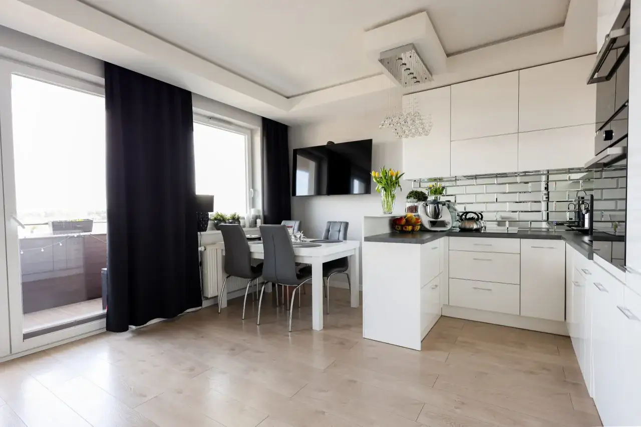 Nowoczesna kuchnia z jadalnią, gdzie pakiety wykończenia pod klucz tworzą spójną całość. Białe meble, lustrzana ściana i elegancka lampa.