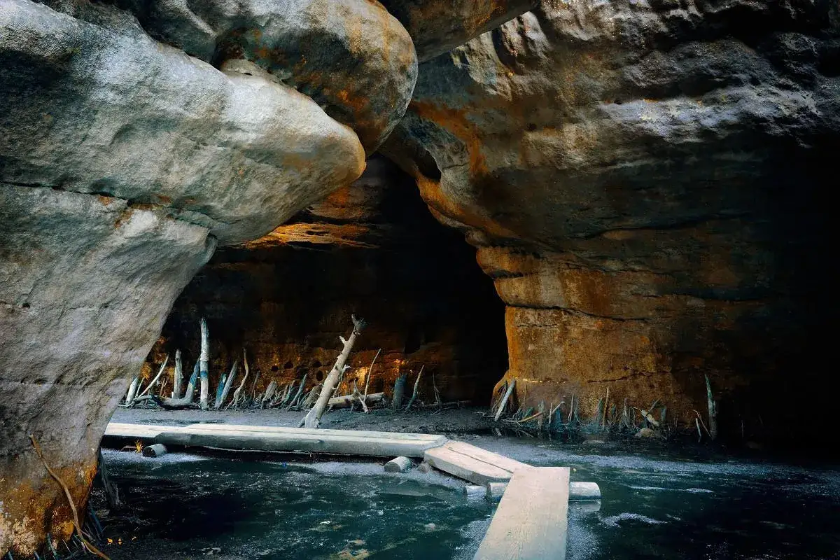 Wąska ścieżka z desek prowadzi przez wodę do jaskini. Wokół Błędnych Skał, w mroku, widać korzenie i powalone drzewa.