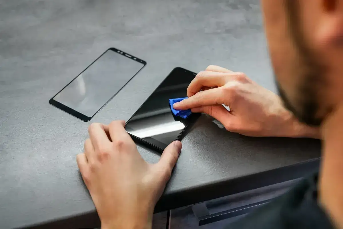 Osoba czyści ekran telefonu przed naklejaniem szkła ochronnego. Obok leży nowe szkło.