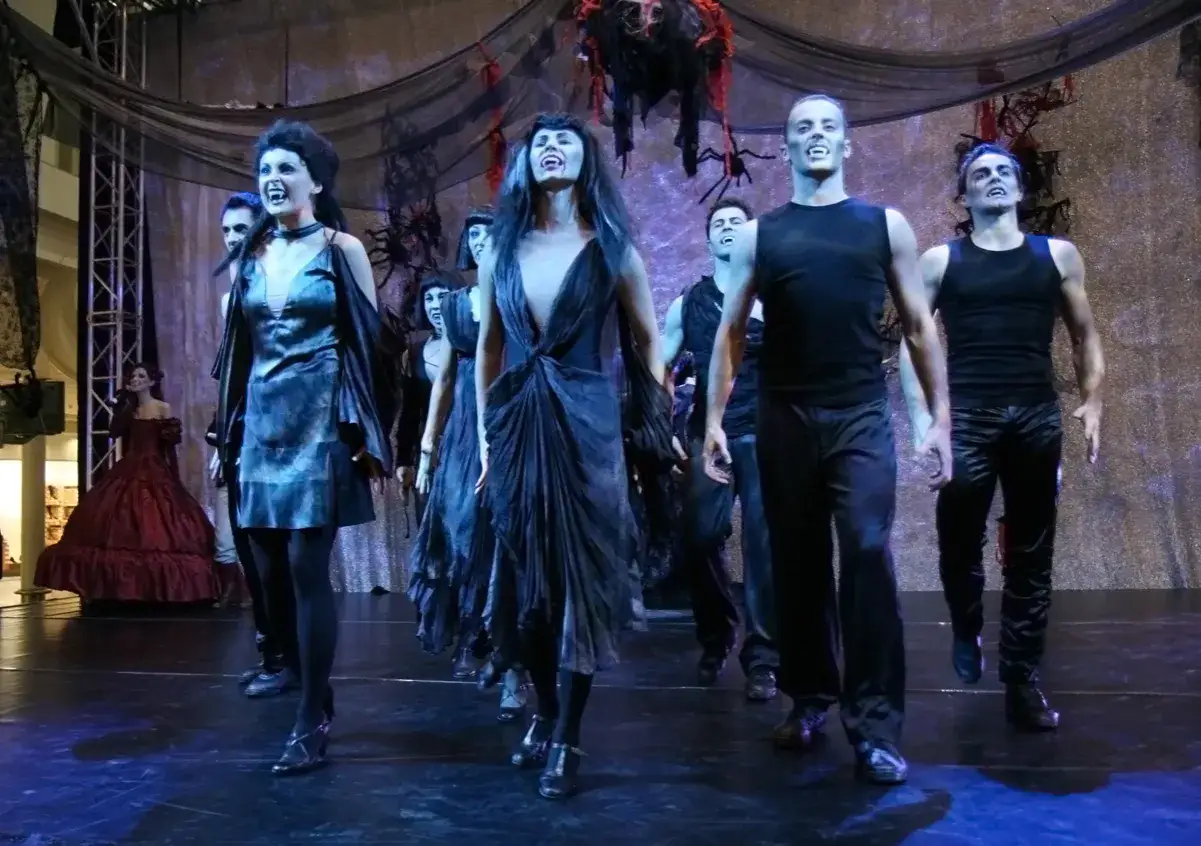 Aktorzy w strojach wampirów, z wyciągniętymi rękami i otwartymi ustami, tańczą na scenie. Taniec wampirów musical w pełnej krasie.