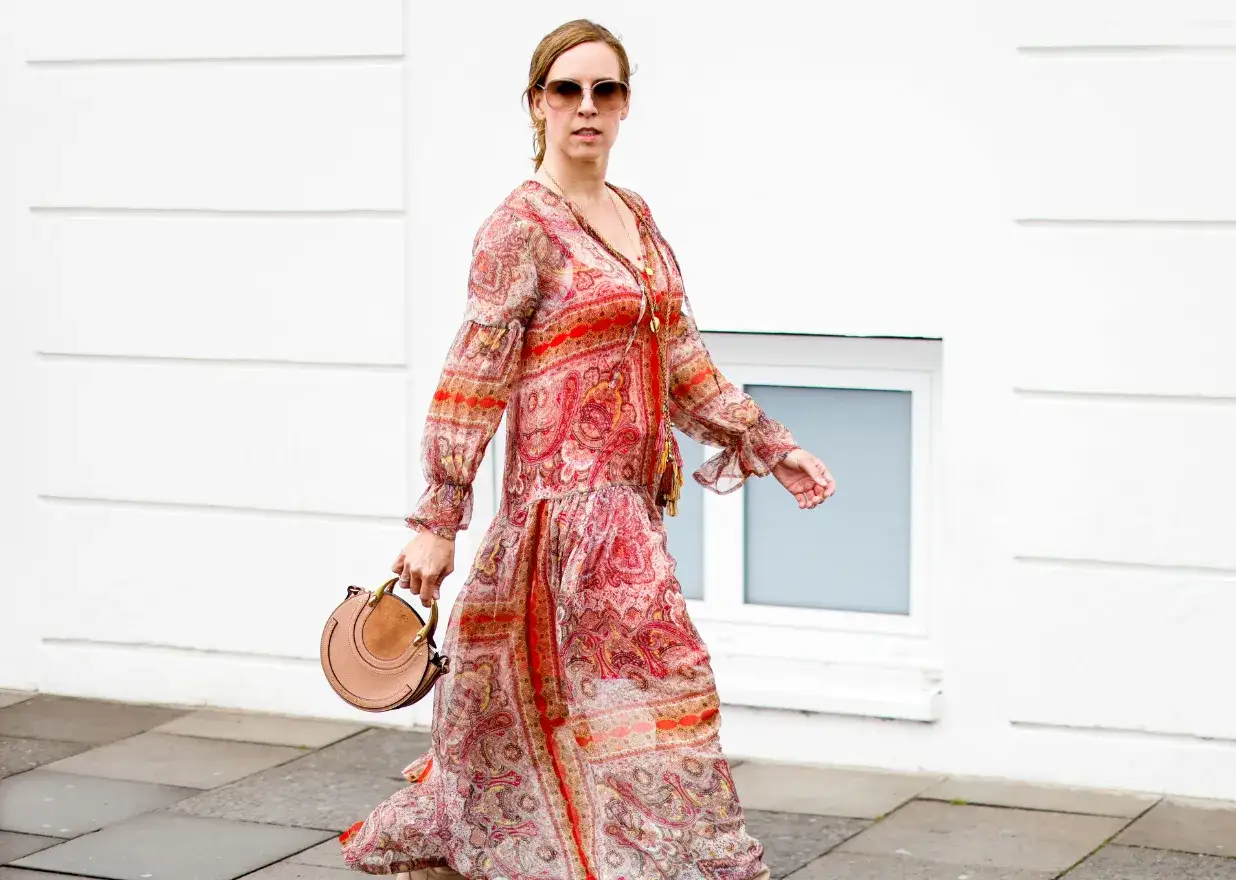 Kobieta w zwiewnej, wzorzystej czerwonej sukience boho z frędzlami i okrągłą torebką na spacerze.