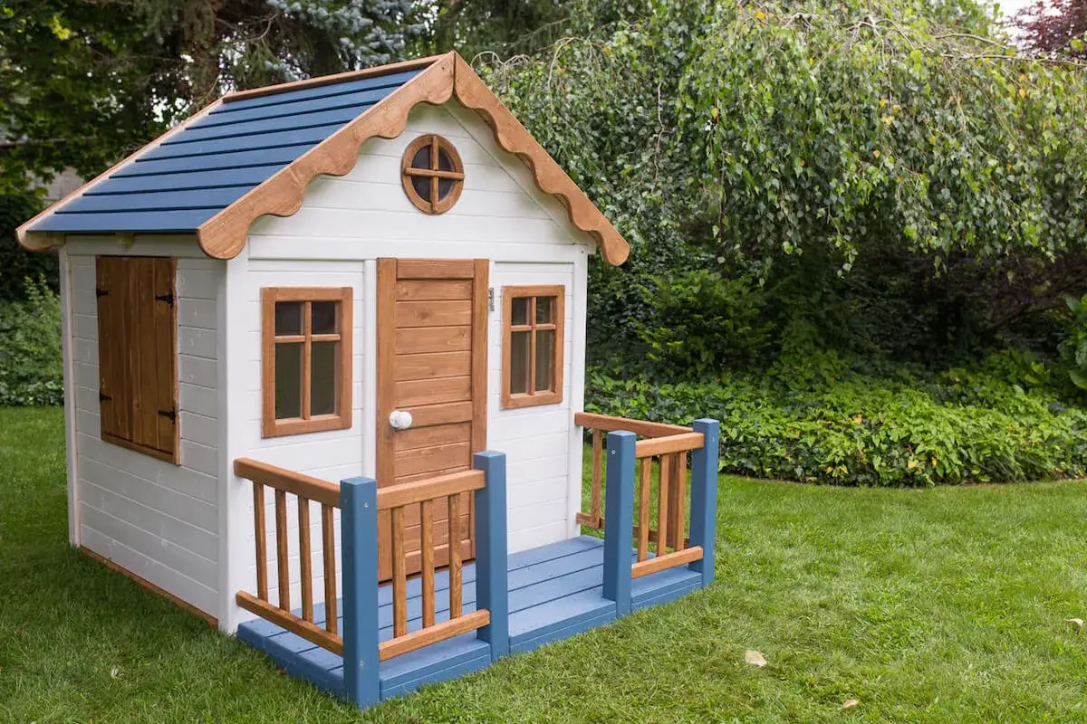 Jak pomalować domek dla dzieci? Biały domek z niebieskim dachem i drewnianymi elementami stoi na trawniku.