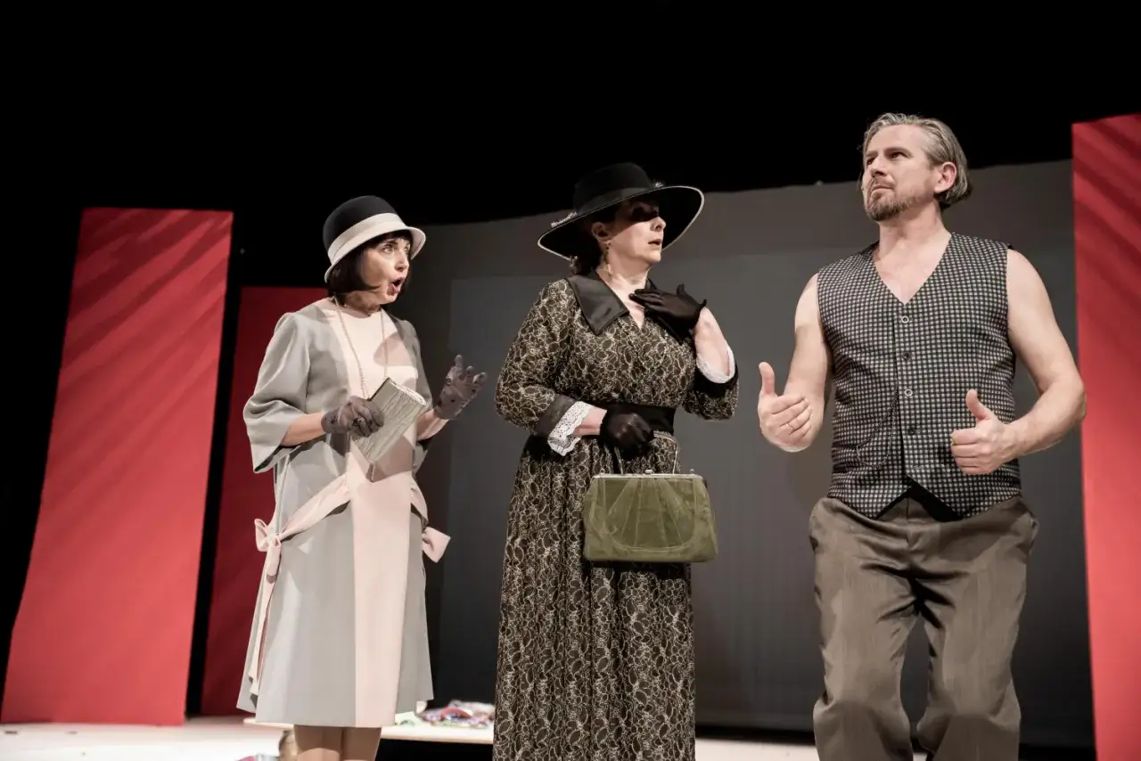 Aktorzy na scenie, jedna kobieta w szarej sukience, druga w czarnej, a obok nich mężczyzna w kamizelce. Może to scena z 