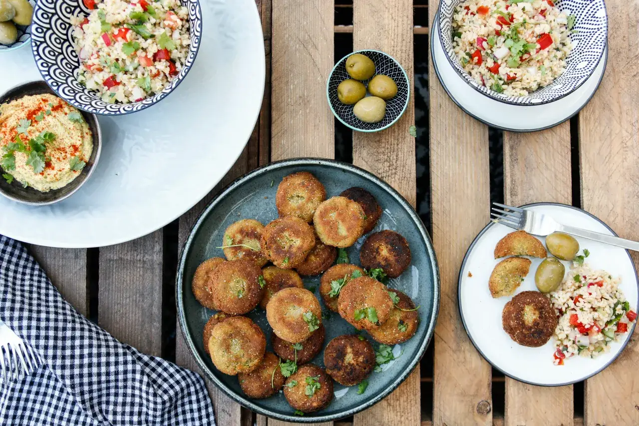 Falafel kontra kotlety sojowe na drewnianym stole. Obok miseczki z hummusem, sałatką tabbouleh i oliwkami.