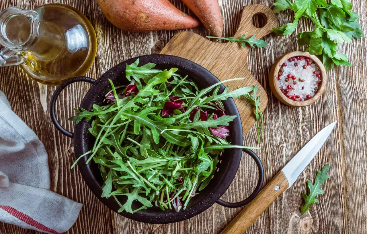 Świeża rukola w misce, obok bataty, oliwa i przyprawy. Dowiedz się, co zrobić, żeby rukola nie była gorzka.