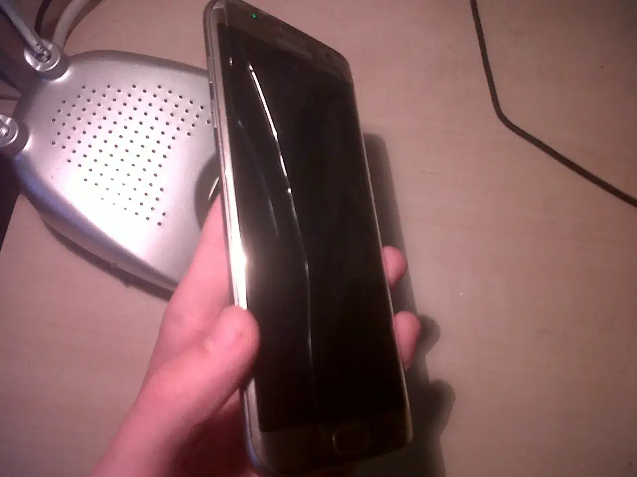 Dłoń trzyma telefon z pękniętym szkłem hartowanym. Obok leży srebrna, perforowana podstawka.