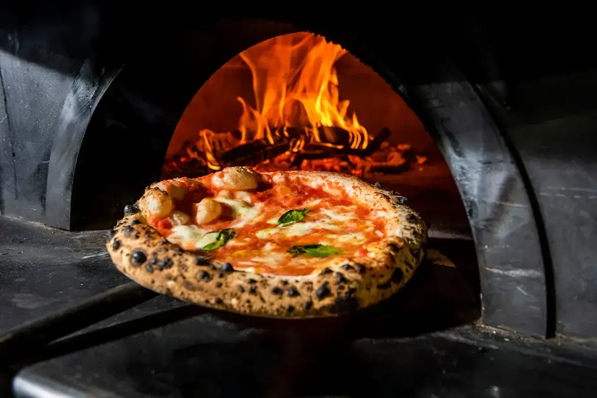 Piec opala się ogniem, a obok niego leży pizza, przypominając o tym, gdzie powstała pizza.