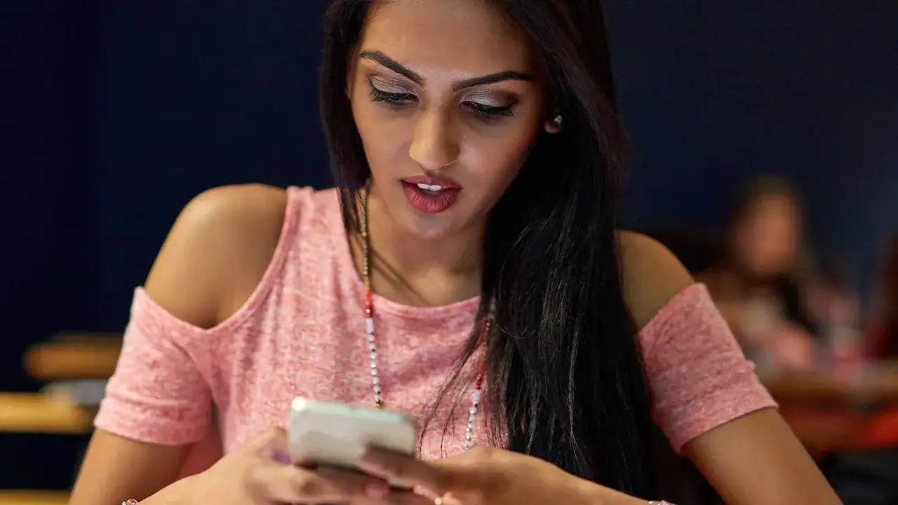 Dziewczyna z długimi włosami, w r&oacute;żowej bluzce, przegląda telefon. Zastanawia się, co napisać dziewczynie, żeby było jej miło.