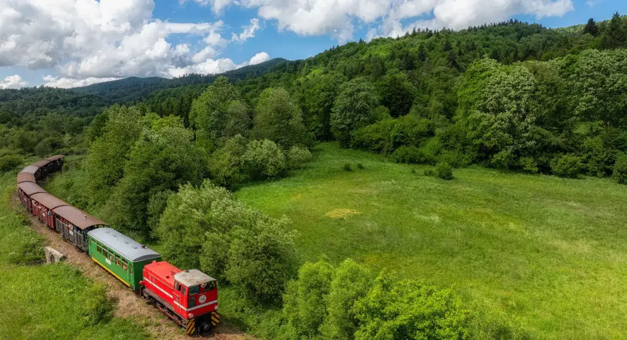 Pociąg z czerwonym lokomotywem i brązowymi wagonami przemierza malownicze tereny, oferując niezwykłe podróże koleją w Polsce.
