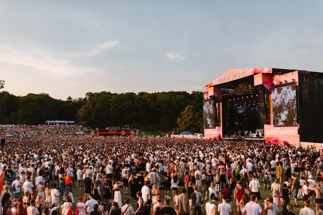 Tłum ludzi na festiwalu Bittersweet, scena z neonowym napisem i ekranami, zach&oacute;d słońca.