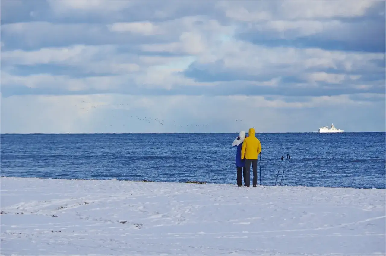 Para spaceruje po zaśnieżonej plaży, wdychając świeże powietrze. Kiedy jod nad morzem, widok statku i ptaków w oddali dodaje uroku zimowemu krajobrazowi.