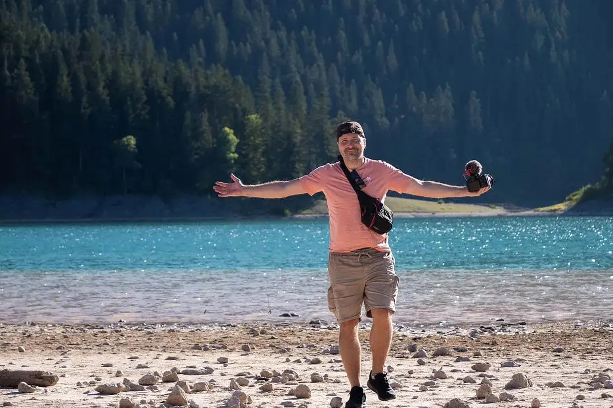 Mężczyzna z aparatem na brzegu turkusowego jeziora w Czarnog&oacute;rze, z rozłożonymi ramionami, jakby chciał powiedzieć: 