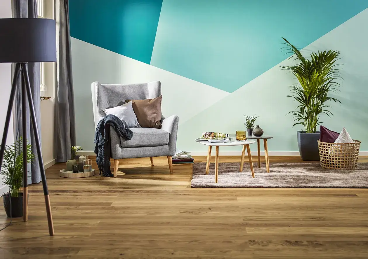 Nowoczesny salon z geometrycznym wzorem na ścianie w odcieniach turkusu i mięty. Wygodny fotel, stoliki kawowe i rośliny tworzą przytulną atmosferę. Świetny pomysł na pomalowanie pokoju.